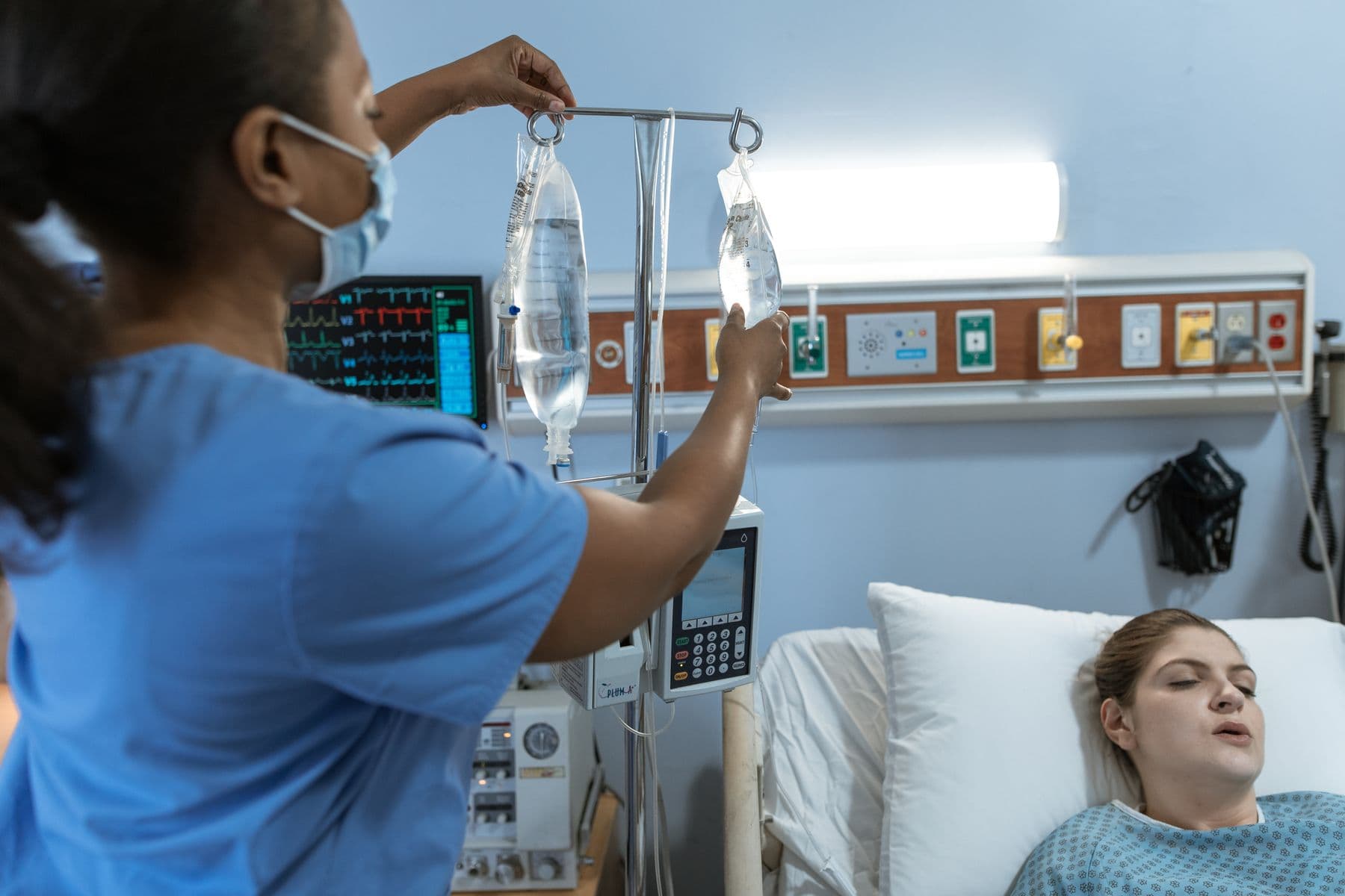 A nurse in blue scrubs hangs an IV bag at a patient's bedside